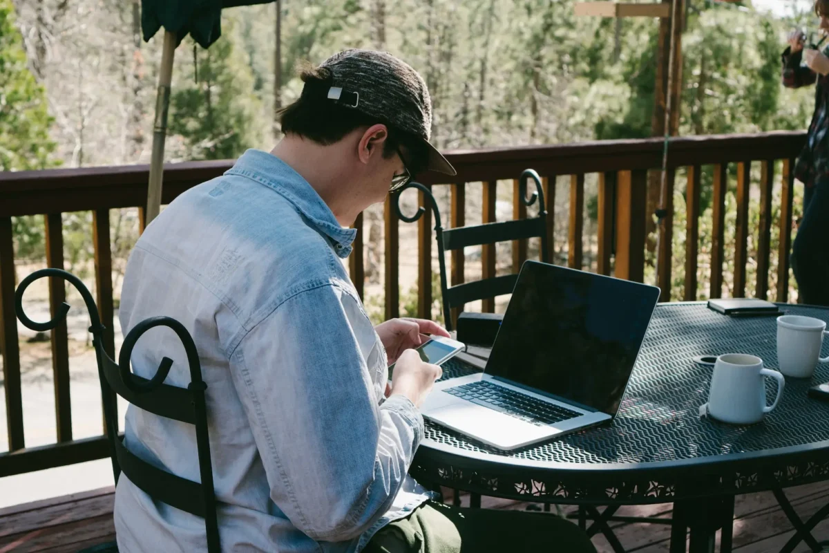 Man in a cap and glasses using a laptop sitting at a metal table.